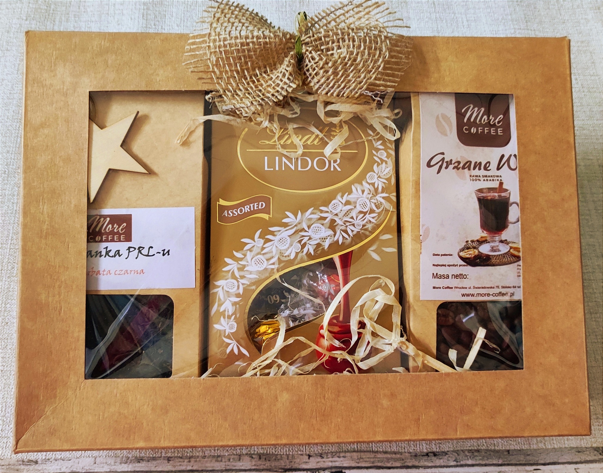 Kartonowy prezent z okienkami, zawierający kawę, czekoladki Lindor i drewnianą gwiazdkę, przewiązany jutowym sznurkiem z kokardą.