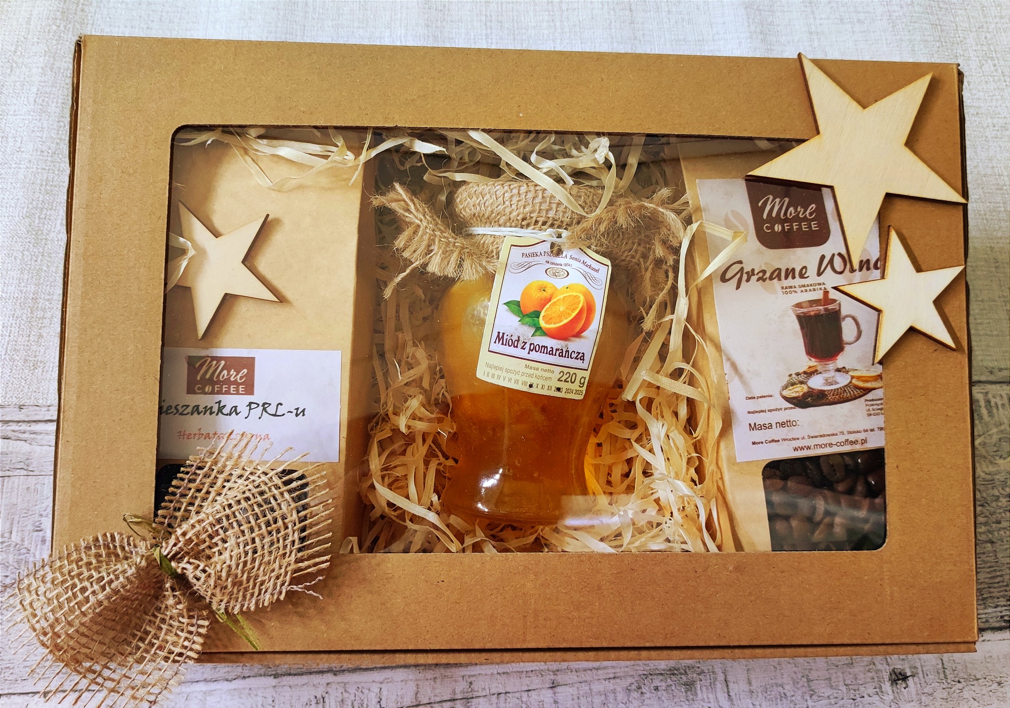 Kartonowe pudełko z okienkiem prezentujące miód pomarańczowy, kawę grzane wino oraz herbatę PRL-u, udekorowane jutowym sznurkiem i drewnianymi gwiazdkami.