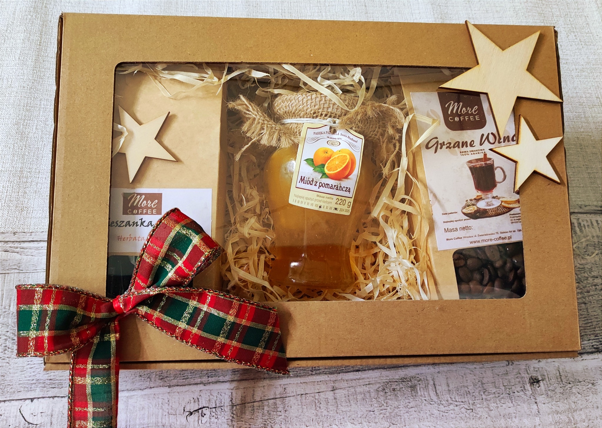 Ozdobny kartonowy zestaw prezentowy z okienkiem, wypełniony miodem pomarańczowym, kawą ziarnistą, herbatą i dekoracjami z drewnianych gwiazdek, przewiązany czerwoną wstążką w kratę.