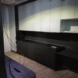 Krzysztof Lubieniecki - Częściowo zamontowana, nowoczesna kuchnia z czarnymi szafkami dolnymi i białymi górnymi, widoczne przewody elektryczne i niedokończone wykończenie sufitu.