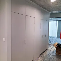 Długa, zabudowana szafa wnękowa z szarymi, gładkimi frontami, zamontowana w biurze, z podłogą zabezpieczoną folią malarską.