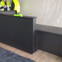 Krzysztof Lubieniecki - Czarna, matowa lada recepcyjna o prostej konstrukcji, ustawiona w pomieszczeniu w trakcie prac wykończeniowych, widoczni pracownicy w odblaskowych kamizelkach. Podłoga zabezpieczona folią malarską.