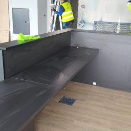 Krzysztof Lubieniecki - Montaż szarej lady recepcyjnej z drewnianym blatem w surowym wnętrzu, widoczni pracownicy w odblaskowych kamizelkach, folia ochronna na podłodze, drabiny i narzędzia w tle.