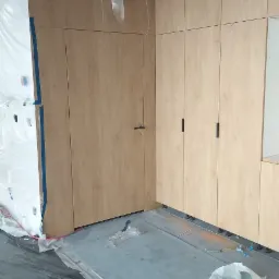 Montaż zabudowy meblowej z drewnianymi frontami, widoczne szafki z czarnymi uchwytami oraz drzwi wkomponowane w zabudowę, podłoga zabezpieczona folią malarską.