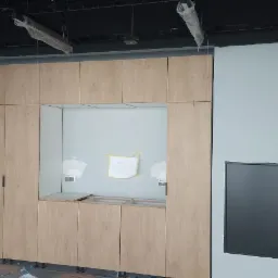 Wnętrze w trakcie wykańczania: zabudowa meblowa z frontami w odcieniach jasnego drewna, widoczne niedokończone podłączenia elektryczne i instalacje pod sufitem, na ścianie przyklejony szkic...