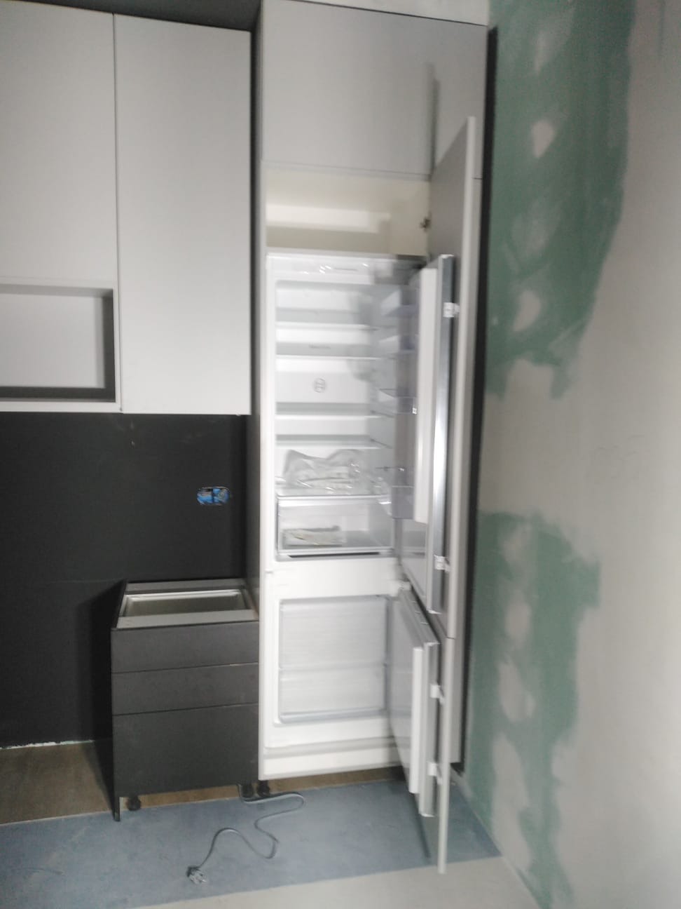 Wnętrze kuchni w trakcie montażu: białe szafki wiszące, czarne szafki dolne, otwarta lodówka w zabudowie, ściana przygotowana do malowania.