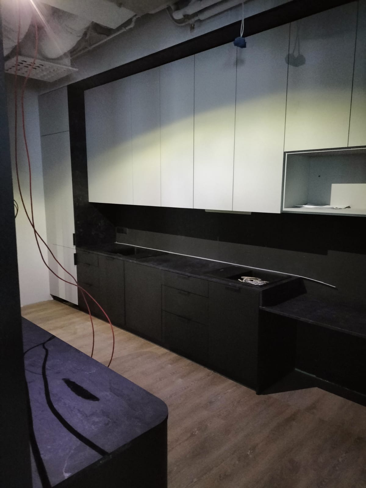 Częściowo zamontowana, nowoczesna kuchnia z czarnymi szafkami dolnymi i białymi górnymi, widoczne przewody elektryczne i niedokończone wykończenie sufitu.