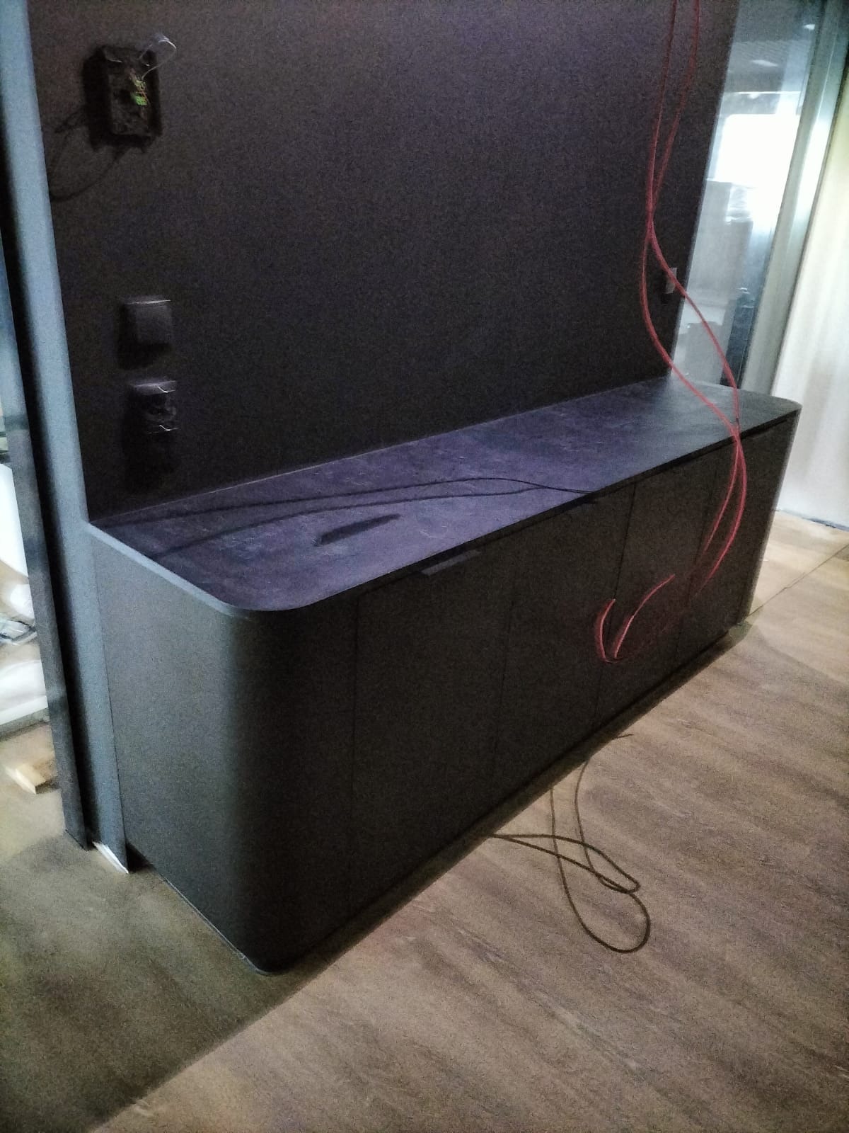 Ciemna szafka RTV z zaokrąglonymi krawędziami i ciemnym blatem, widoczne kable wiszące z tyłu i prowadzone po podłodze, w tle ściana z gniazdkami.