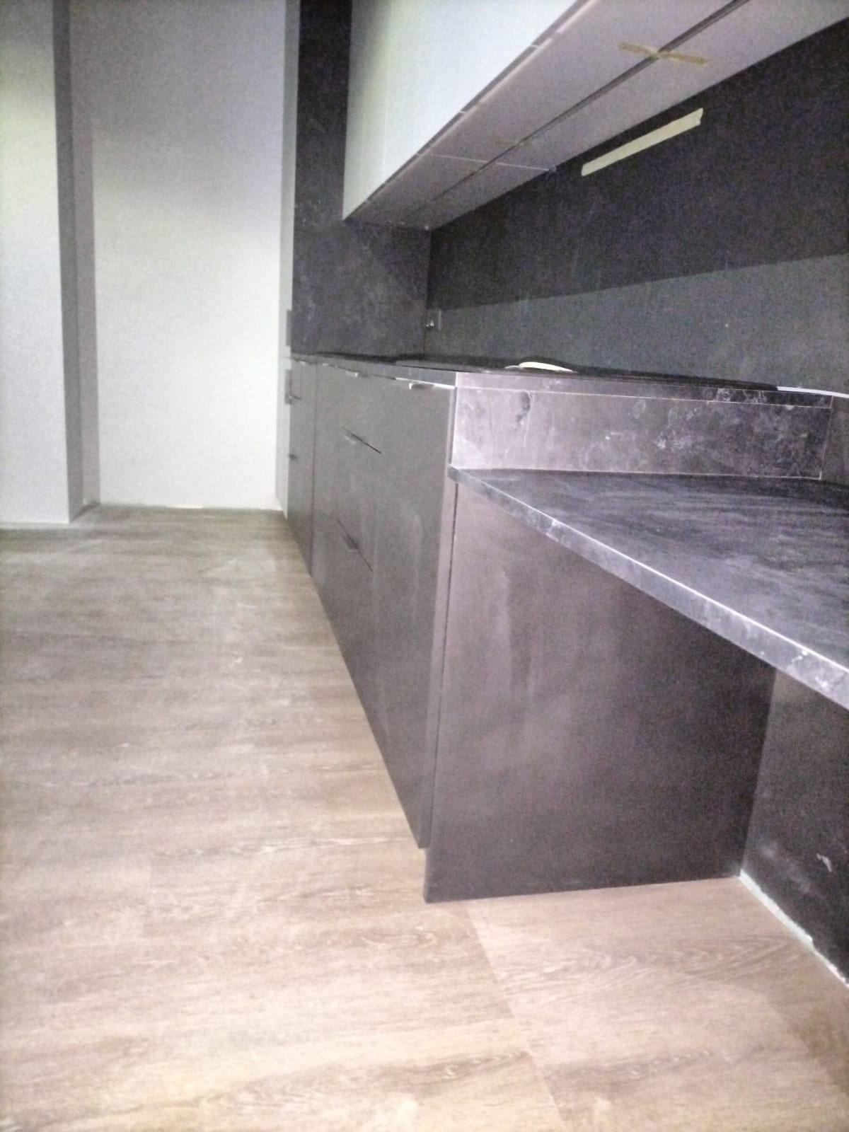 Ciemna zabudowa kuchenna z blatem z kamienia, widok z boku, podłoga wykończona panelami imitującymi drewno.