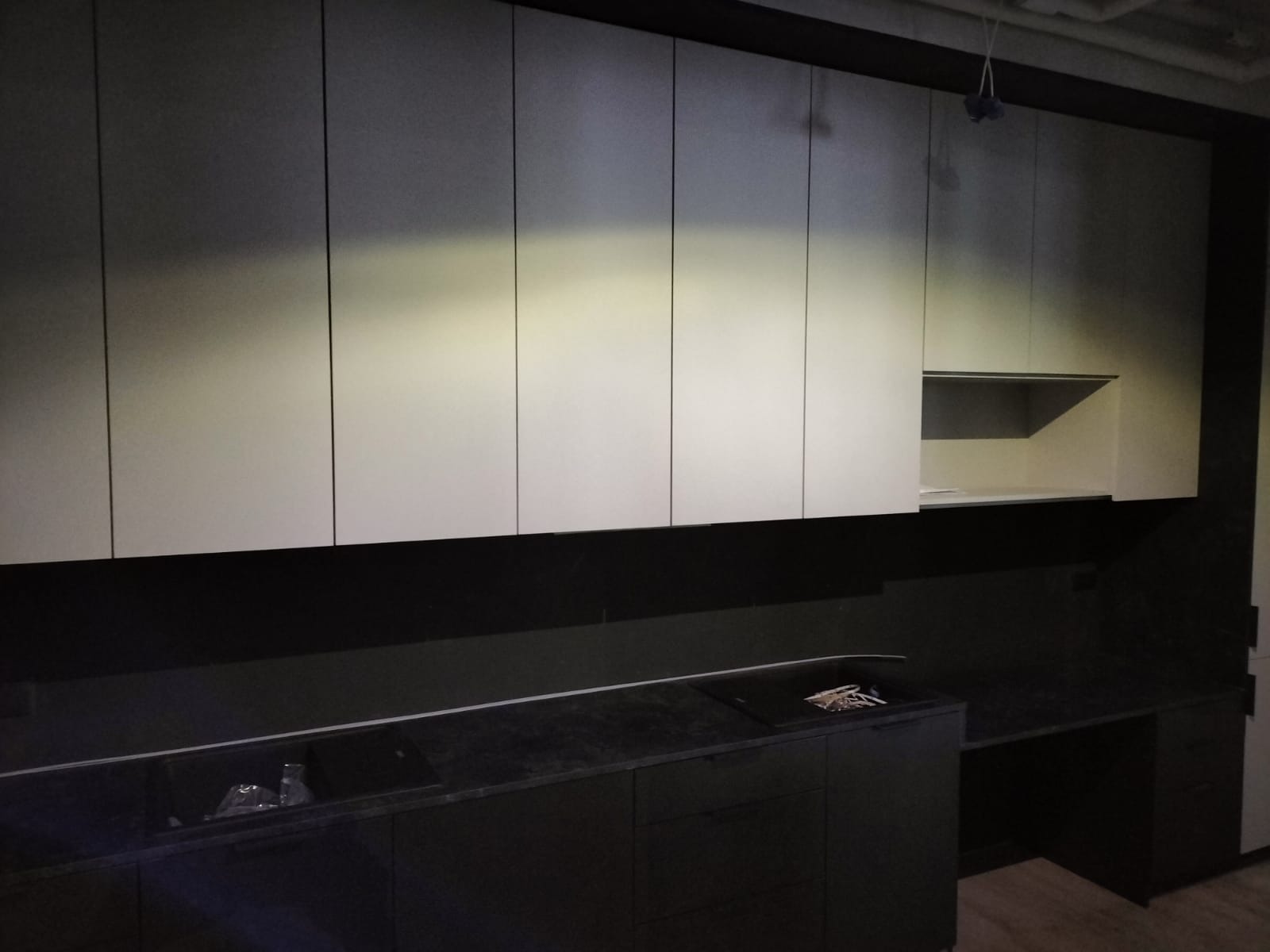 Nowoczesna kuchnia z białymi szafkami górnymi i czarnym blatem, minimalistyczny design, widoczne elementy instalacji elektrycznej pod sufitem.