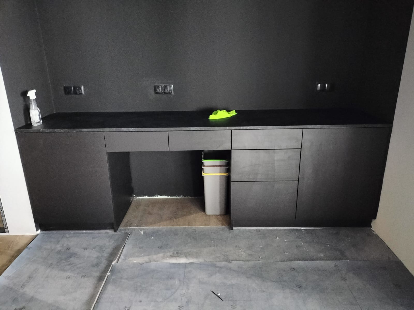 Ciemna zabudowa meblowa z blatem, widoczna podczas montażu na podłodze zabezpieczonej folią, z koszem na śmieci wsuniętym pod blat i butelką środka czyszczącego na blacie.