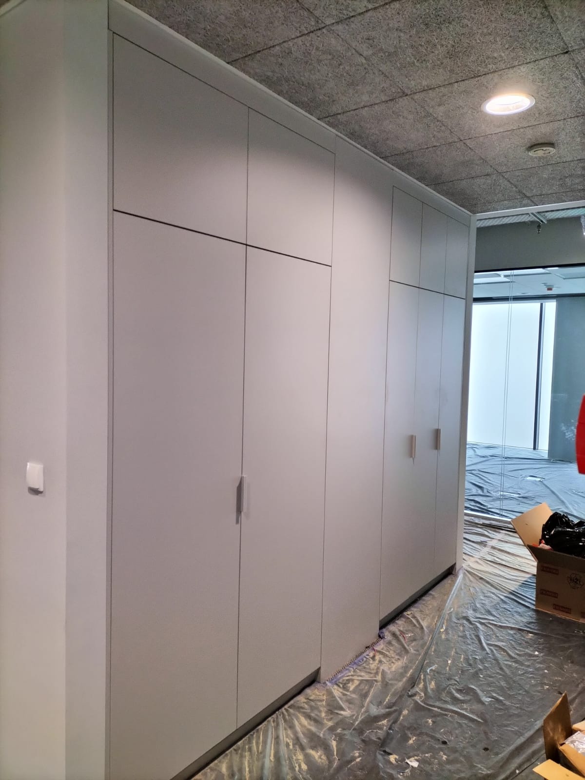 Długa, zabudowana szafa wnękowa z szarymi, gładkimi frontami, zamontowana w biurze, z podłogą zabezpieczoną folią malarską.