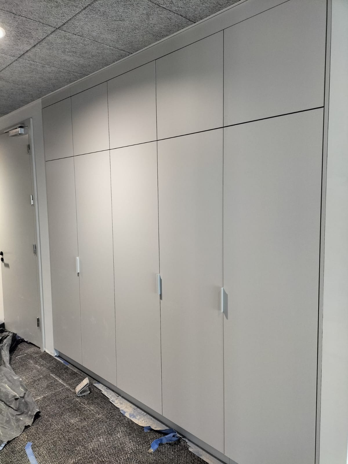 Długa, jasnoszara zabudowa szafy wnękowej z minimalistycznymi uchwytami, widoczna na tle szarej wykładziny i zabezpieczeń podłogi po montażu.