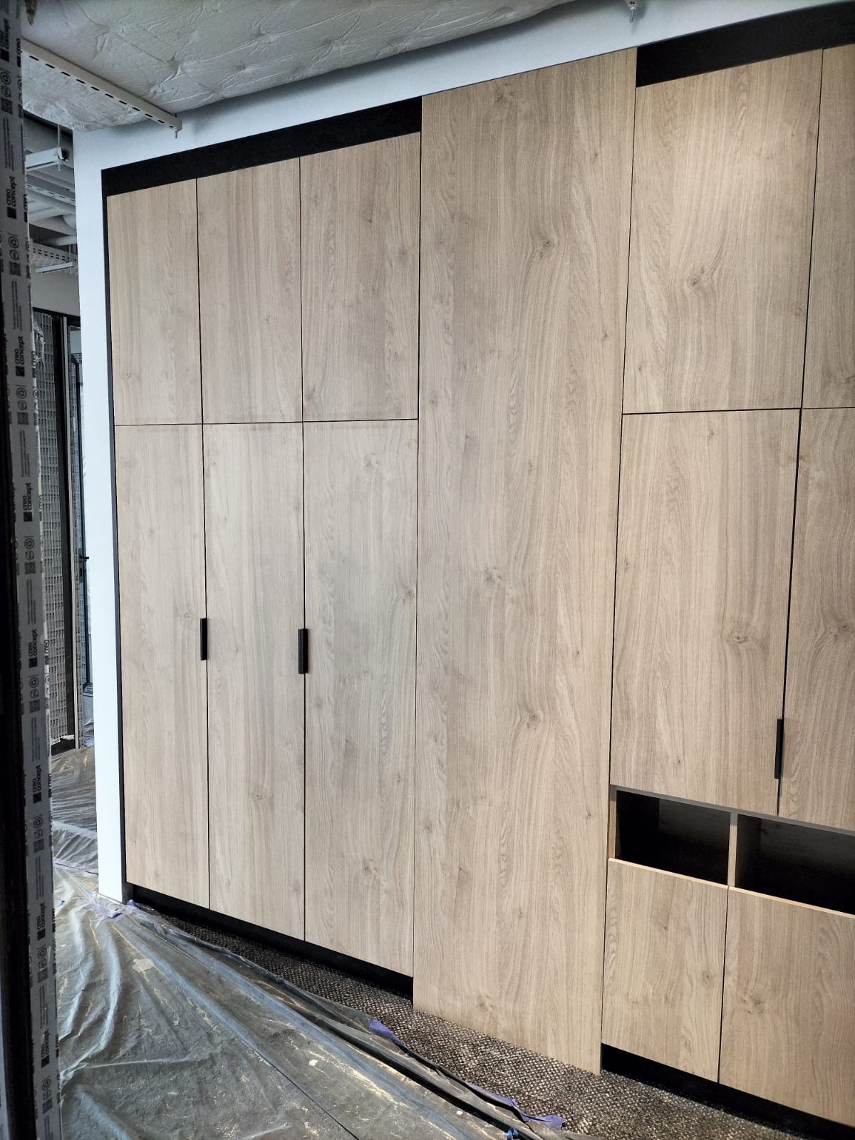 Zabudowa meblowa z drewnopodobnymi frontami i czarnymi detalami, widoczna folia ochronna na podłodze.