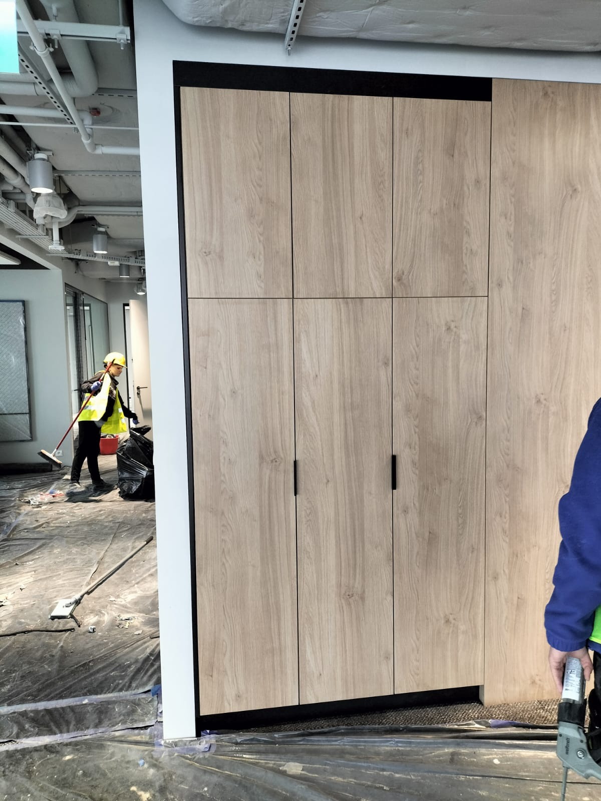 Montaż szafy wnękowej z drewnopodobnymi frontami w biurze w Warcie Bolesławieckiej, widoczna osoba sprzątająca po instalacji.