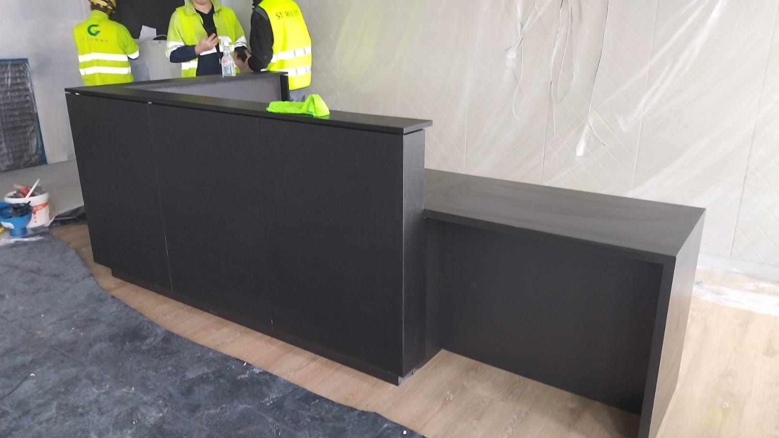 Czarna, matowa lada recepcyjna o prostej konstrukcji, ustawiona w pomieszczeniu w trakcie prac wykończeniowych, widoczni pracownicy w odblaskowych kamizelkach. Podłoga zabezpieczona folią malarską.