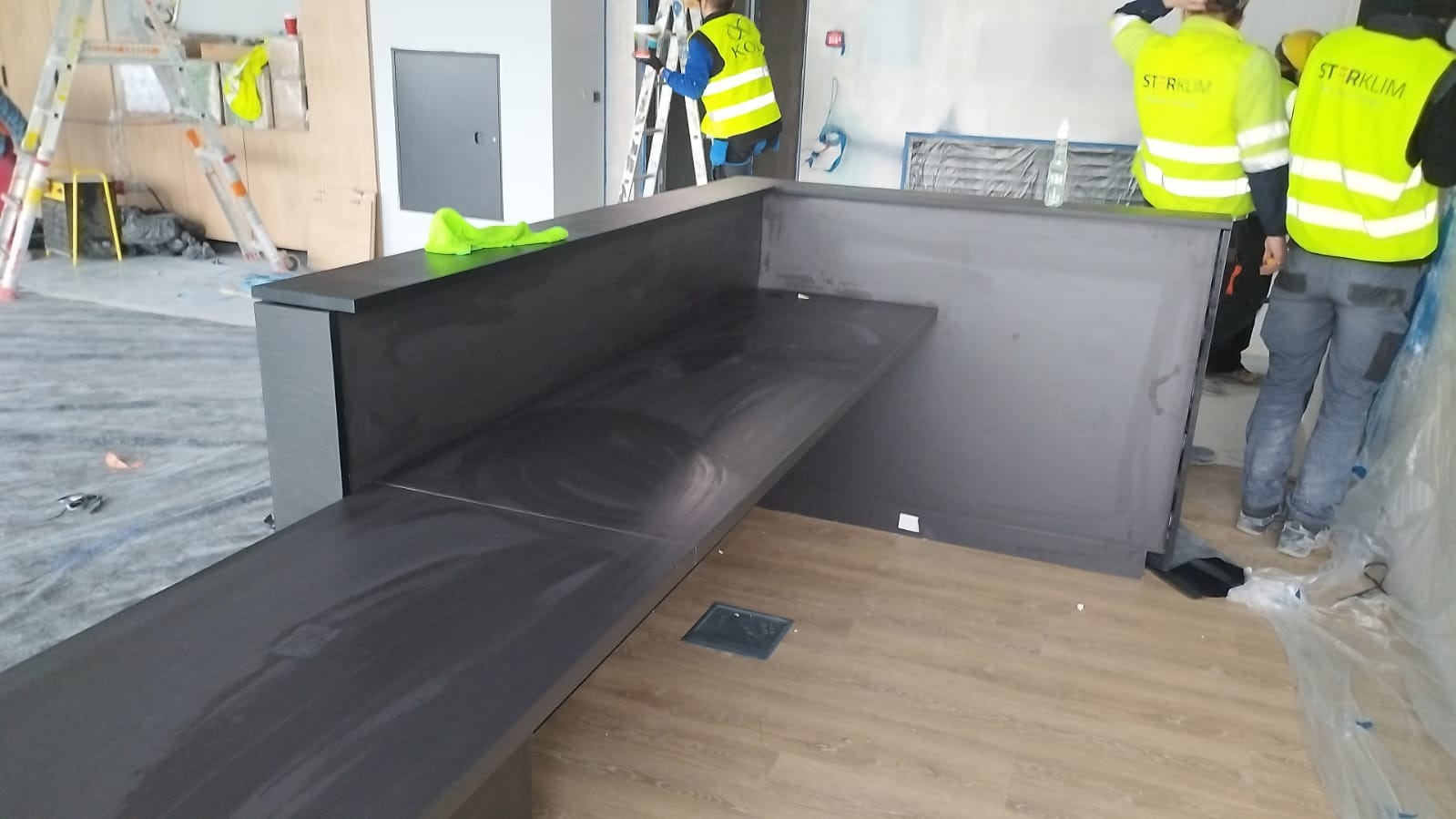 Montaż szarej lady recepcyjnej z drewnianym blatem w surowym wnętrzu, widoczni pracownicy w odblaskowych kamizelkach, folia ochronna na podłodze, drabiny i narzędzia w tle.