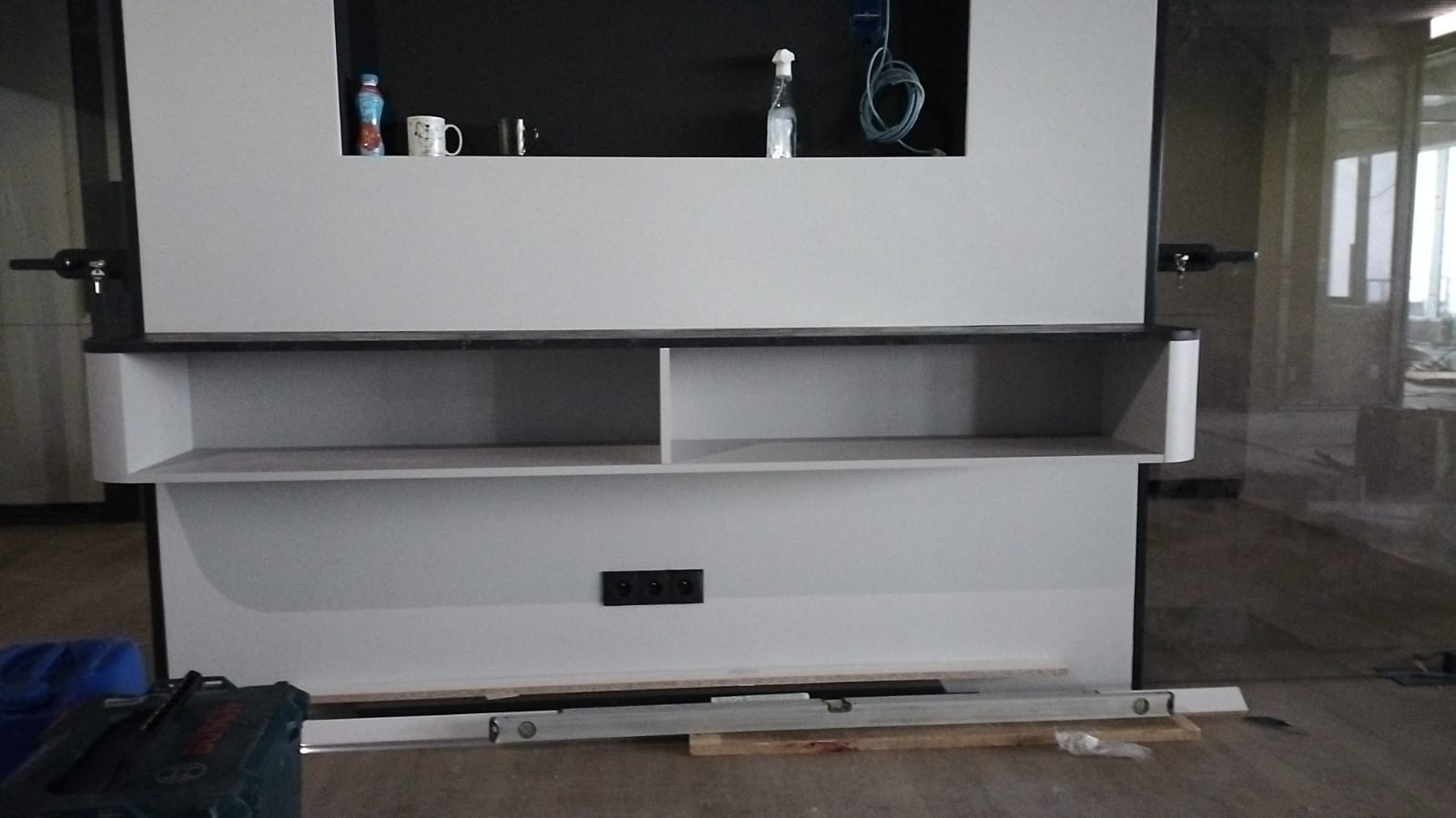 Biała zabudowa meblowa z półkami i czarną wnęką, na której stoją butelka, kubek i dozownik. Na podłodze leżą poziomice i skrzynka narzędziowa.