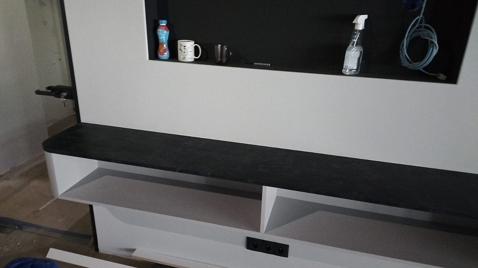 Półka z czarnym blatem i białymi elementami pod spodem, wbudowana w ścianę z czarną wnęką, na której stoją butelka, kubek i spryskiwacz.