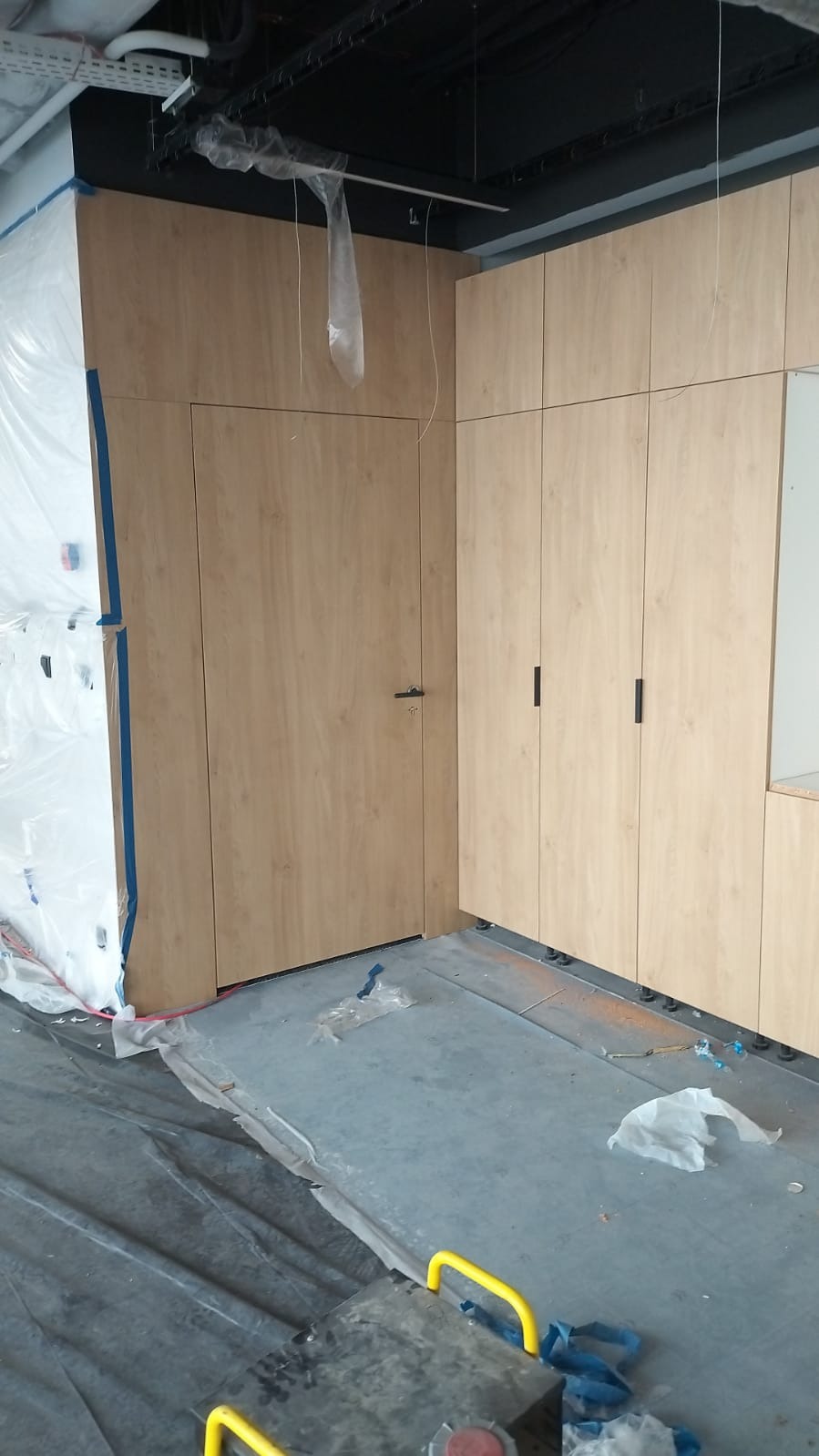 Montaż zabudowy meblowej z drewnianymi frontami, widoczne szafki z czarnymi uchwytami oraz drzwi wkomponowane w zabudowę, podłoga zabezpieczona folią malarską.