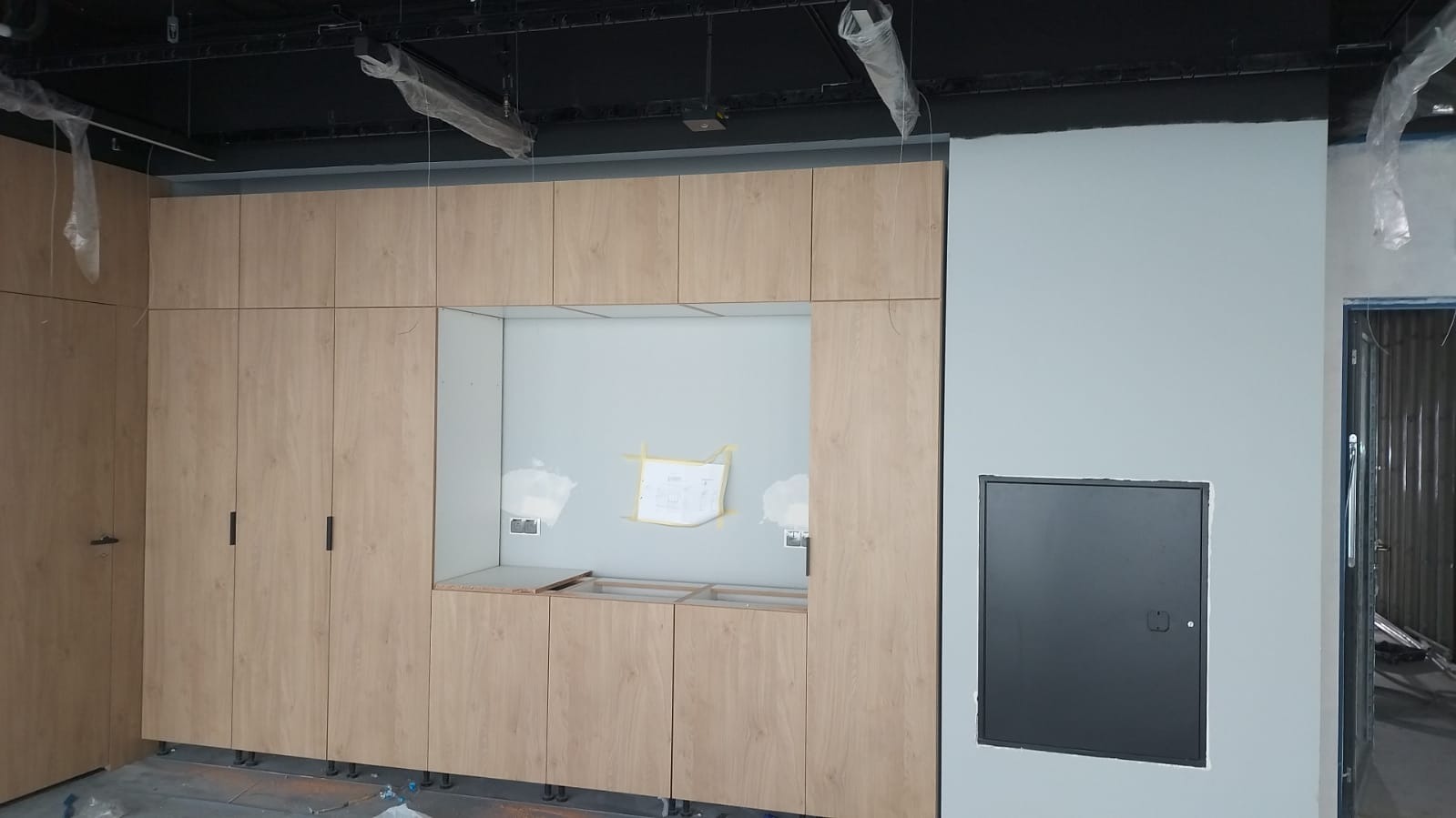 Wnętrze w trakcie wykańczania: zabudowa meblowa z frontami w odcieniach jasnego drewna, widoczne niedokończone podłączenia elektryczne i instalacje pod sufitem, na ścianie przyklejony szkic...