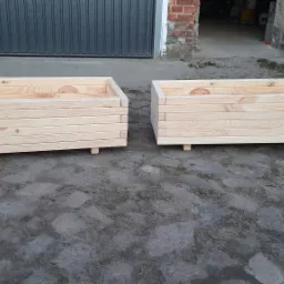 Dwa surowe, drewniane kwietniki skrzynkowe z jasnego drewna, ustawione na zewnątrz na szarym bruku, z widocznymi słojami drewna i małymi nóżkami, w tle brama garażowa i fragment muru.