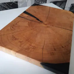 Kwadratowy blat z drewna z wyraźnym rysunkiem słojów, zalany czarną żywicą na krawędziach i w pęknięciach, leżący na białym stole.