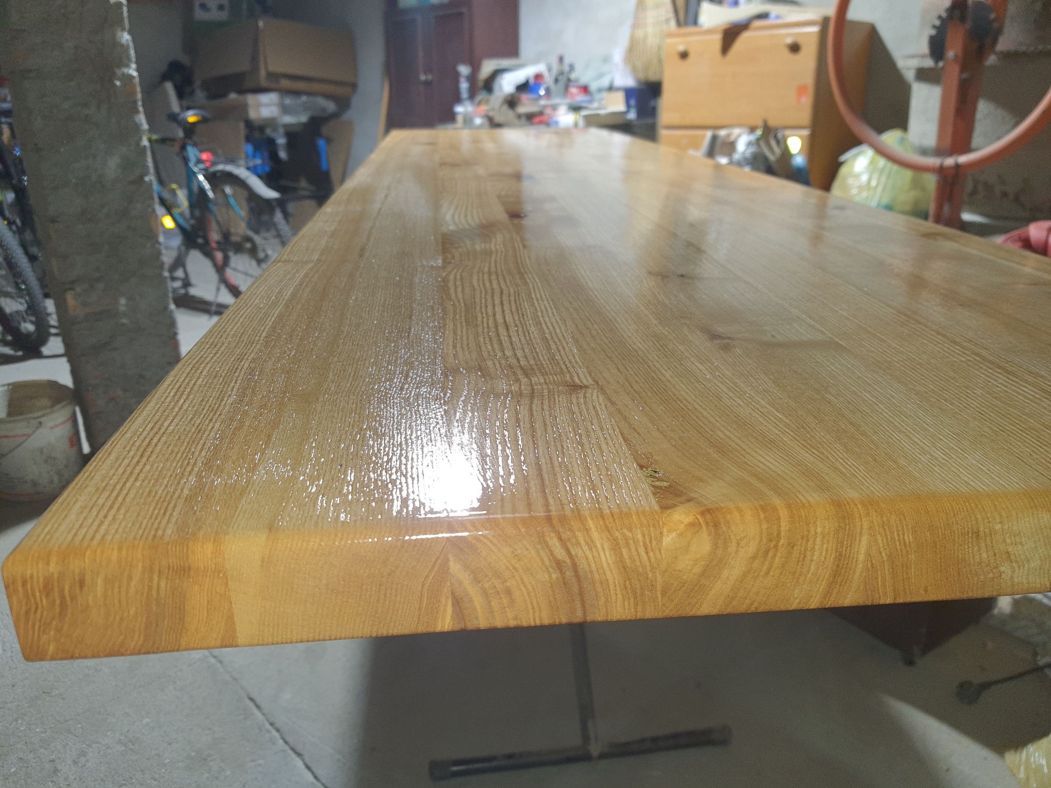 Lakierowany blat drewniany na metalowych nogach, widoczna struktura drewna, w tle rower i przedmioty w garażu.