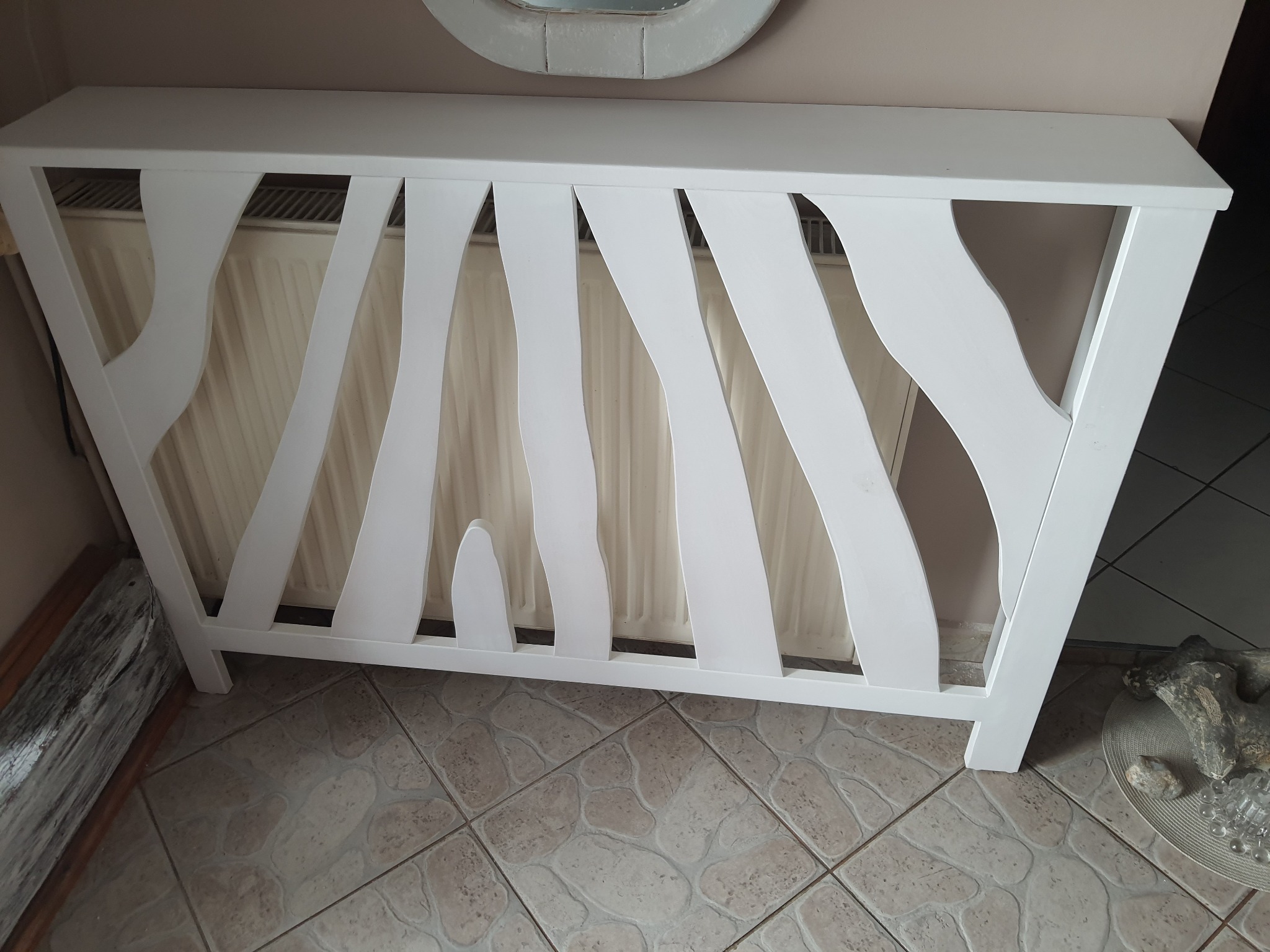 Biała osłona na grzejnik z pionowymi wycięciami w kształcie fal, stojąca na kafelkach, z dekoracyjnymi elementami obok.