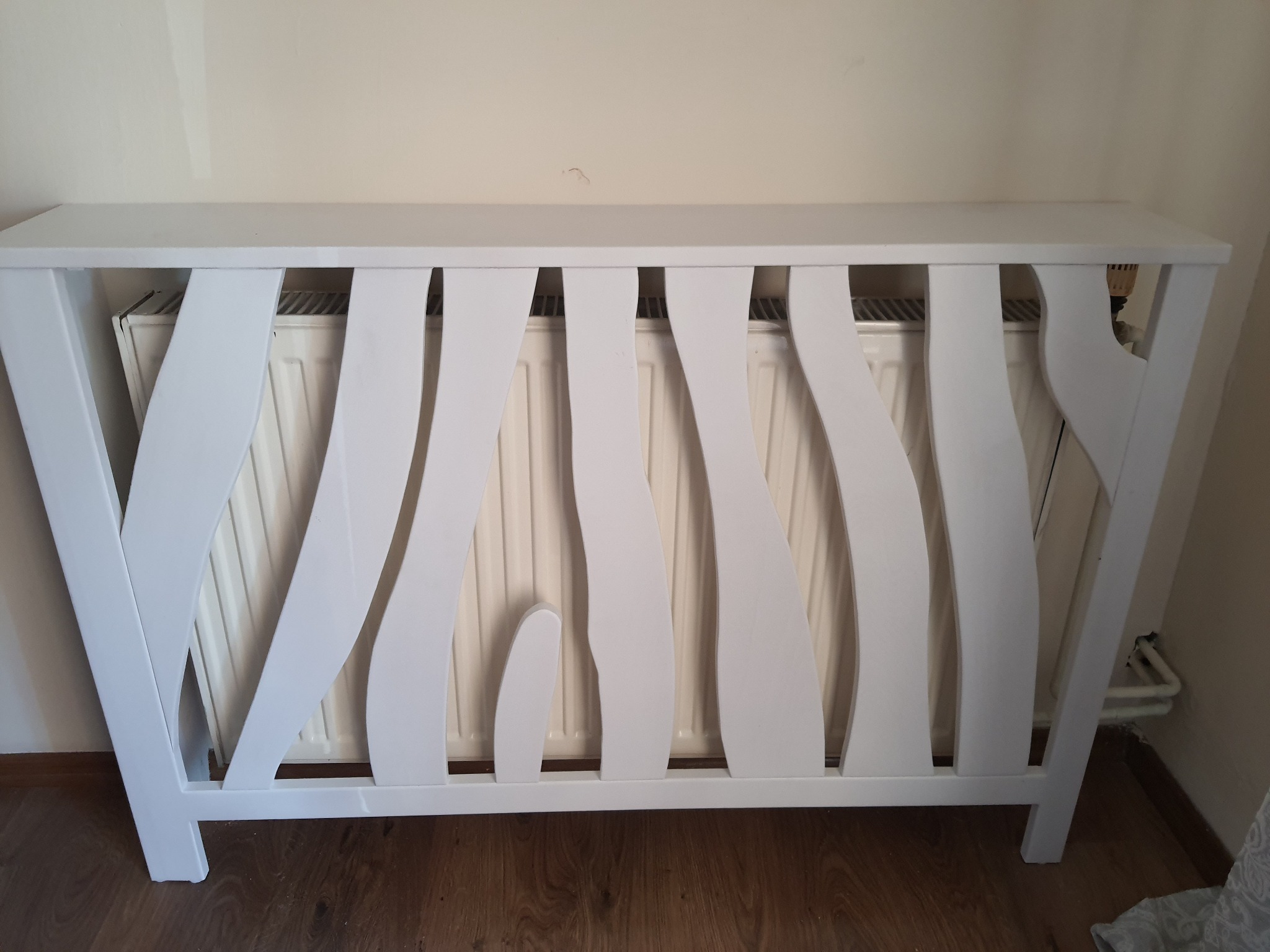 Biała, drewniana osłona dekoracyjna na grzejnik z pionowymi, zaokrąglonymi szczeblami w kształcie płomieni, ustawiona przy beżowej ścianie.