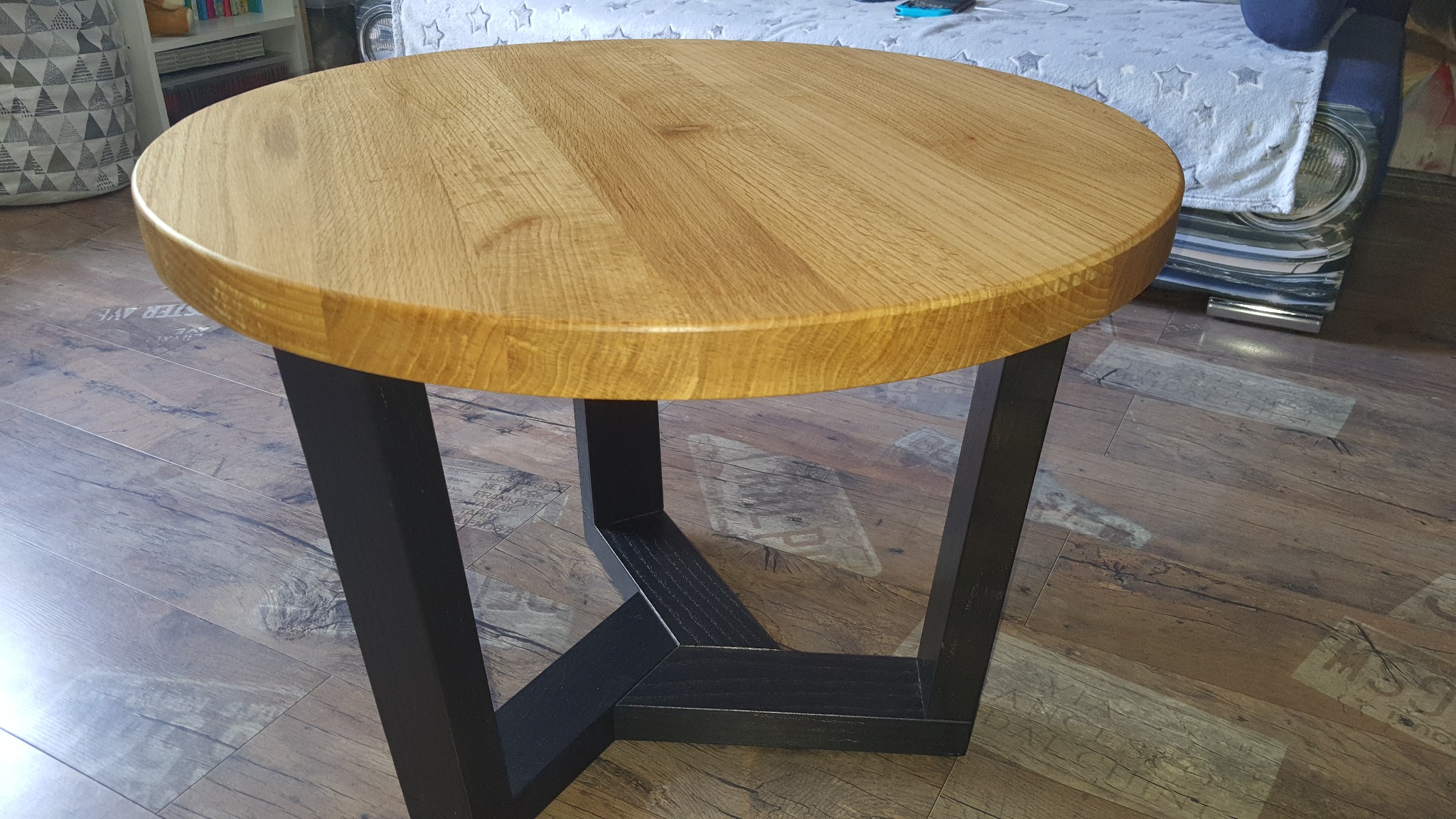 Okrągły stolik kawowy z jasnego drewna z czarnymi, geometrycznymi nogami, ustawiony na panelach podłogowych imitujących drewno.