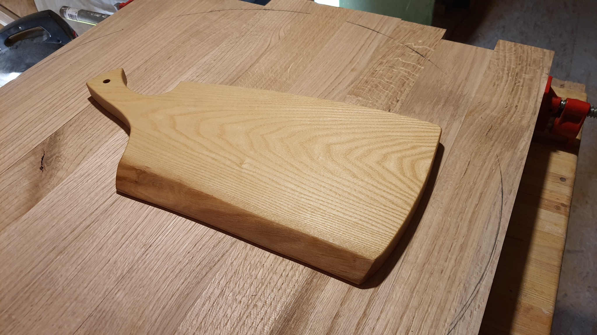 Drewniana deska do krojenia z uchwytem leży na blacie z widocznym zarysem okręgu, obok czerwony ścisk stolarski.