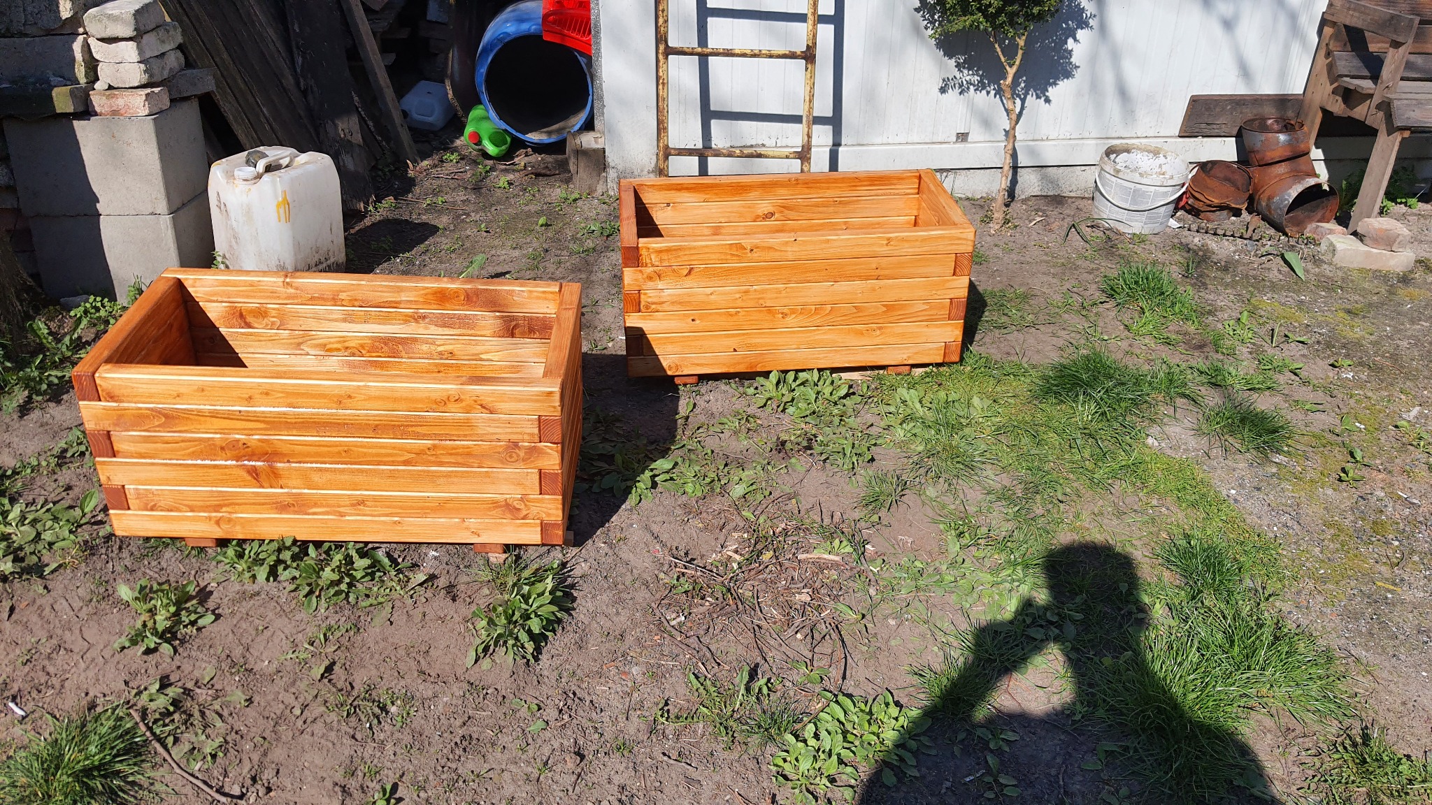 Dwie świeżo zrobione, drewniane skrzynie na rośliny, wykonane z poziomych desek, stoją na trawie w ogrodzie, w tle drabina i inne przedmioty.
