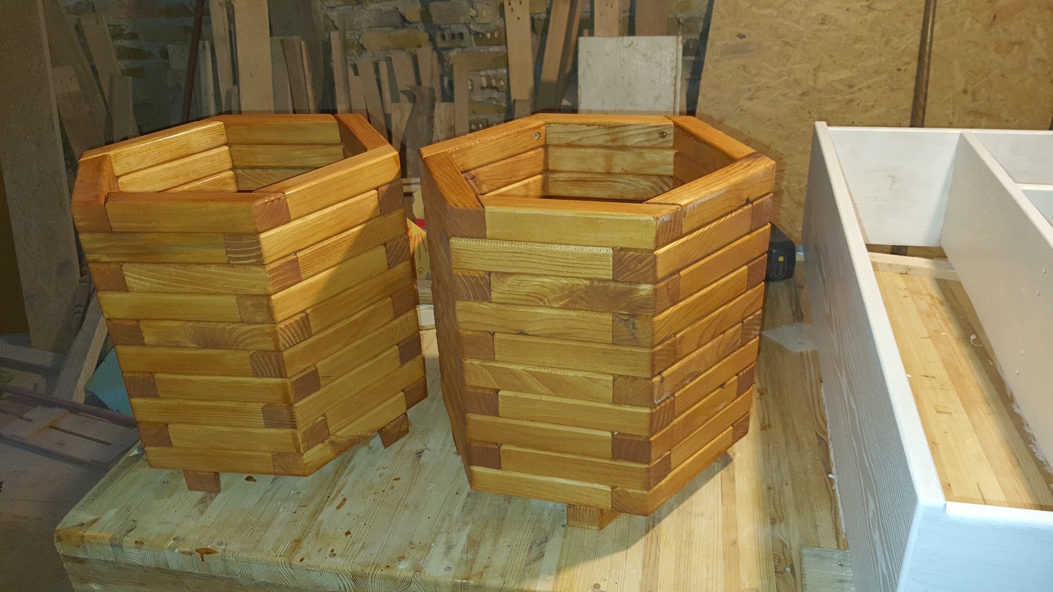 Dwa sześciokątne kwietniki drewniane, wykonane z listew, stoją obok niedokończonego białego regału na tle warsztatu z widocznymi deskami i ceglaną ścianą.