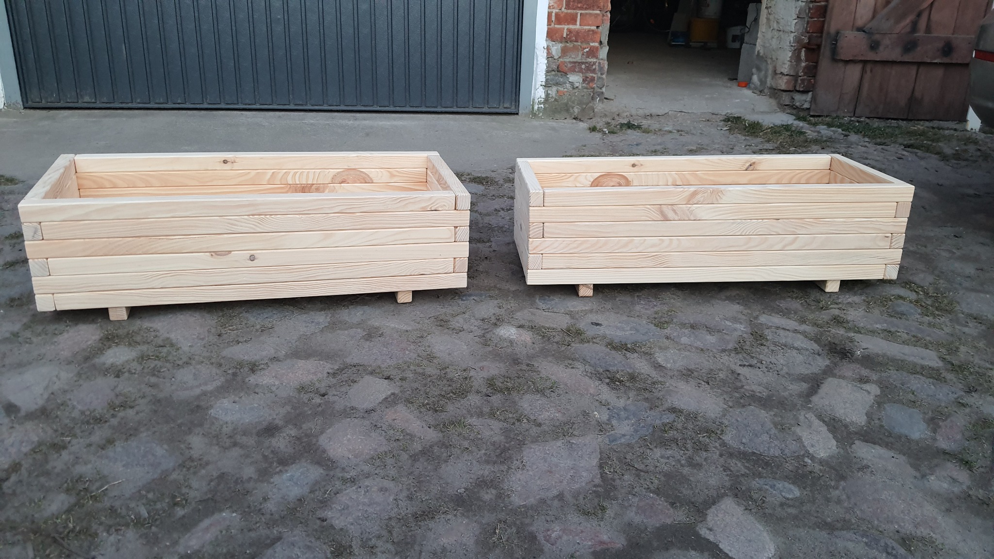 Dwa surowe, drewniane kwietniki skrzynkowe z jasnego drewna, ustawione na zewnątrz na szarym bruku, z widocznymi słojami drewna i małymi nóżkami, w tle brama garażowa i fragment muru.