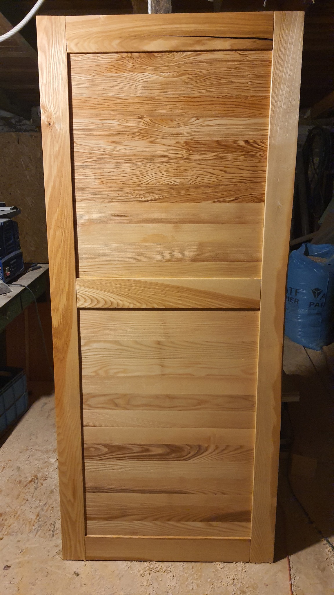 Surowe drewniane drzwi z widocznym usłojeniem, w warsztacie, z workiem na śmieci w tle.