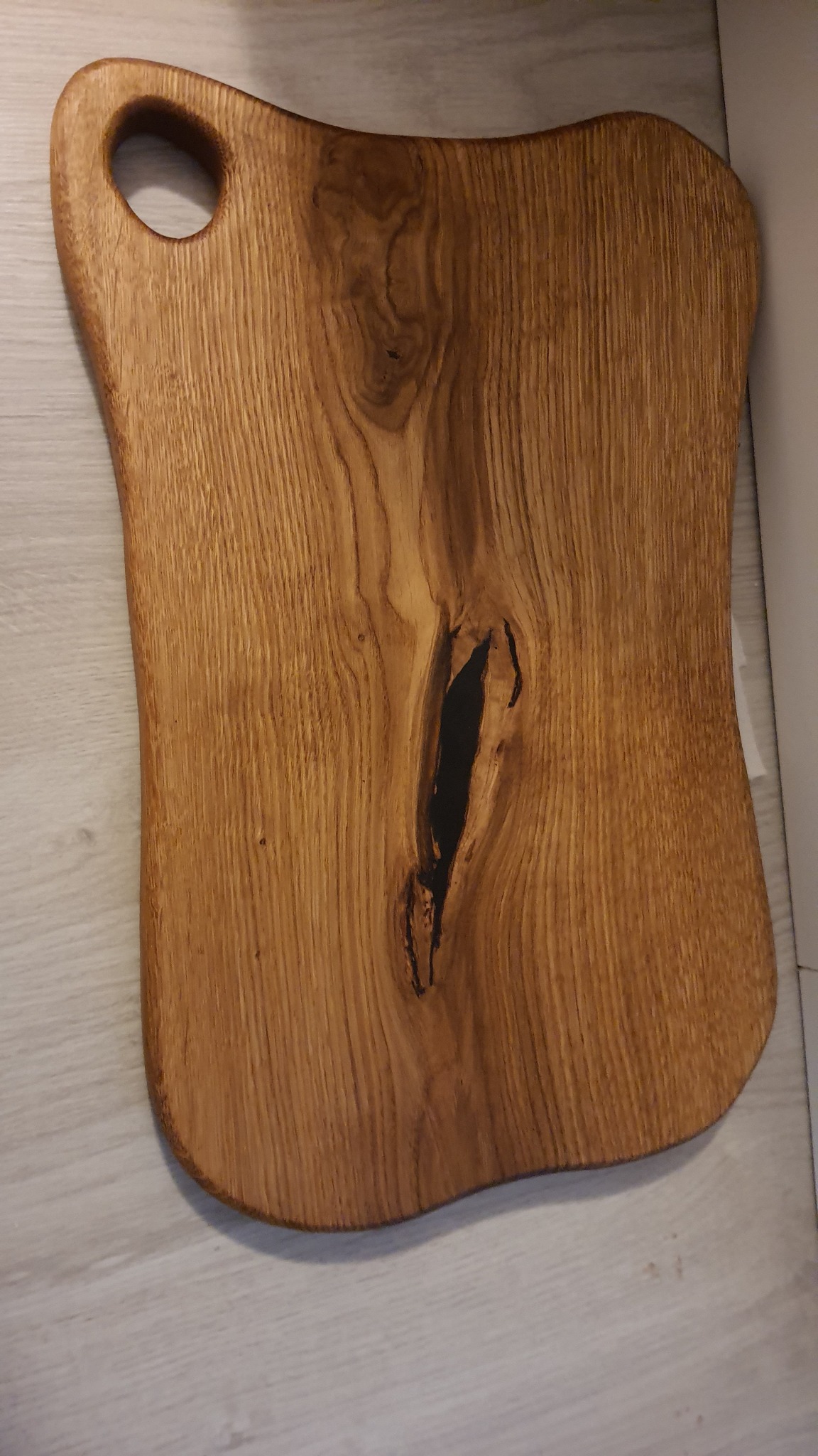 Deska do krojenia z drewna o nieregularnym kształcie z otworem do zawieszenia, z widocznym usłojeniem i naturalną skazą wypełnioną czarną żywicą.