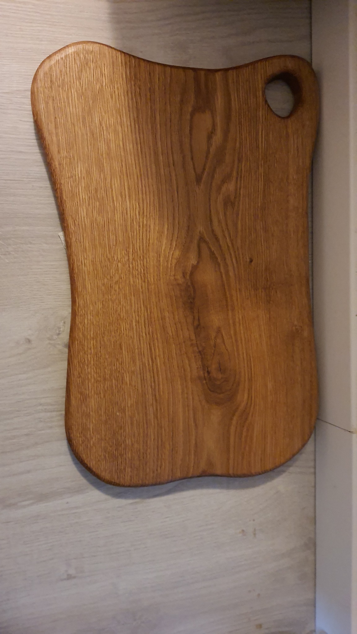 Drewniana deska do krojenia o nieregularnym kształcie z otworem do zawieszenia, leżąca na podłodze imitującej drewno.