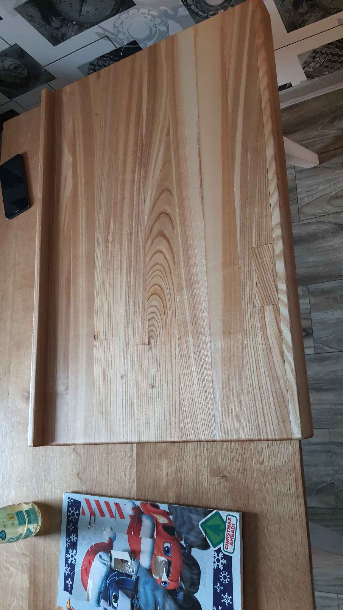 Surowy, jasny blat z litego drewna z widocznymi słojami, leżący na innym blacie, obok leży smartfon i kalendarz adwentowy.