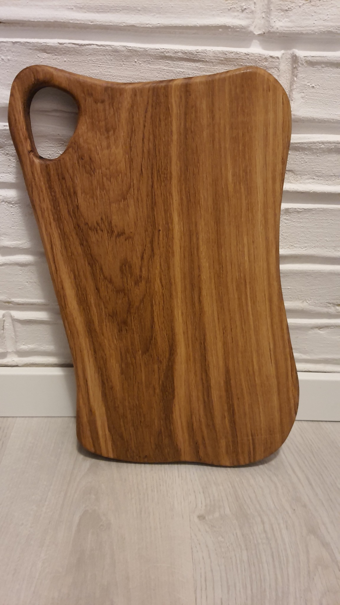 Deska do krojenia z jasnego drewna o nieregularnym kształcie z uchwytem, oparta o ścianę z białej cegły