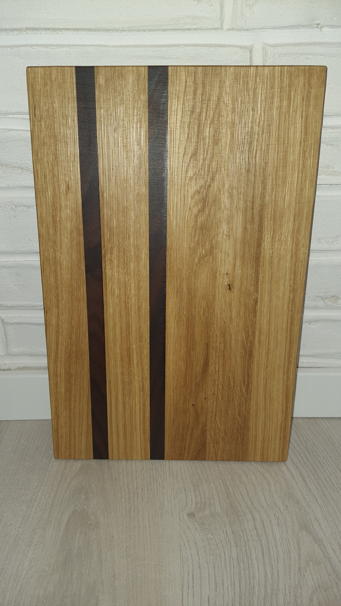 Drewniany blat z trzema wstawkami z ciemniejszego drewna, ustawiony pionowo na podłodze, tło stanowi biała ściana z cegieł.