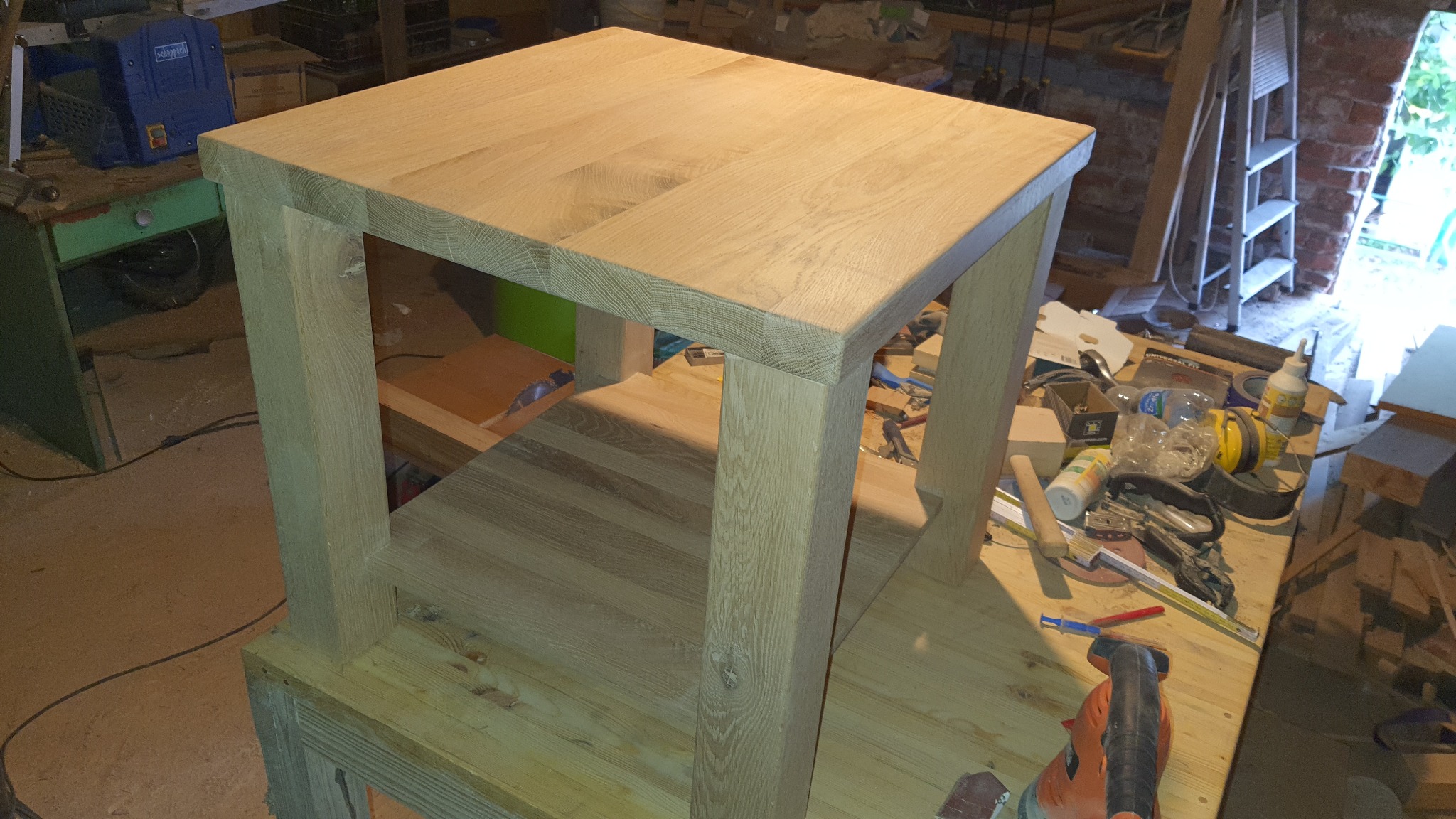 Drewniany stolik kawowy z półką, wykonany z jasnego drewna, widoczny w warsztacie stolarskim z narzędziami i materiałami w tle.