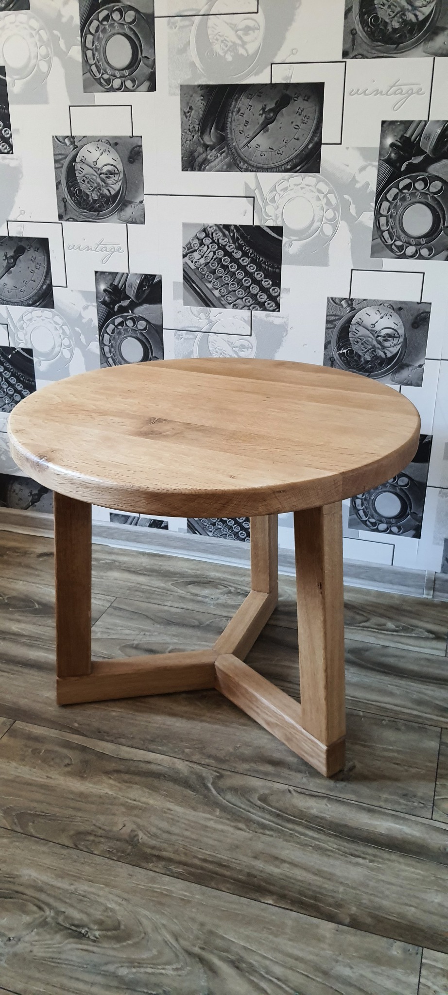 Okrągły, drewniany stolik kawowy z jasnego drewna, z nogami w kształcie litery X, na tle ściany z motywem retro telefonów i maszyn do pisania.