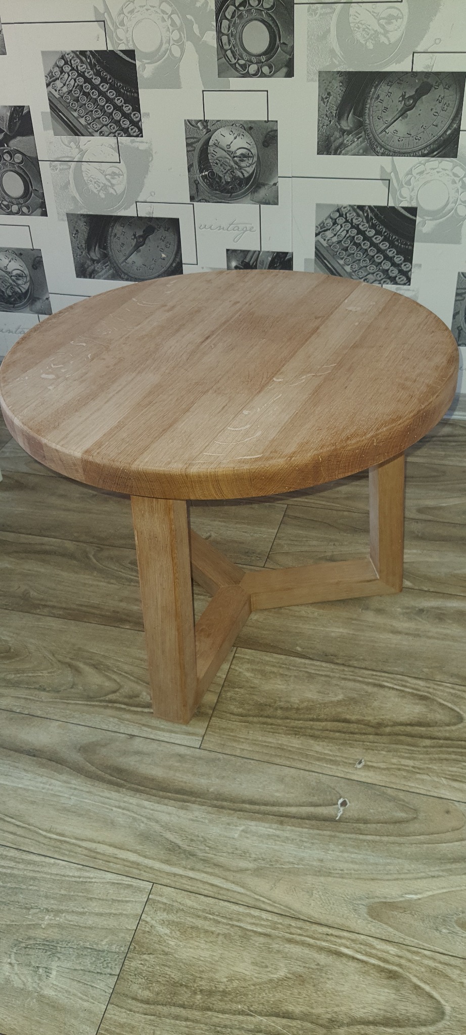 Okrągły stolik kawowy z jasnego drewna z nogami w kształcie trójkąta, na podłodze imitującej drewno, z tapetą w tle z motywem zegarów i maszyn do pisania.