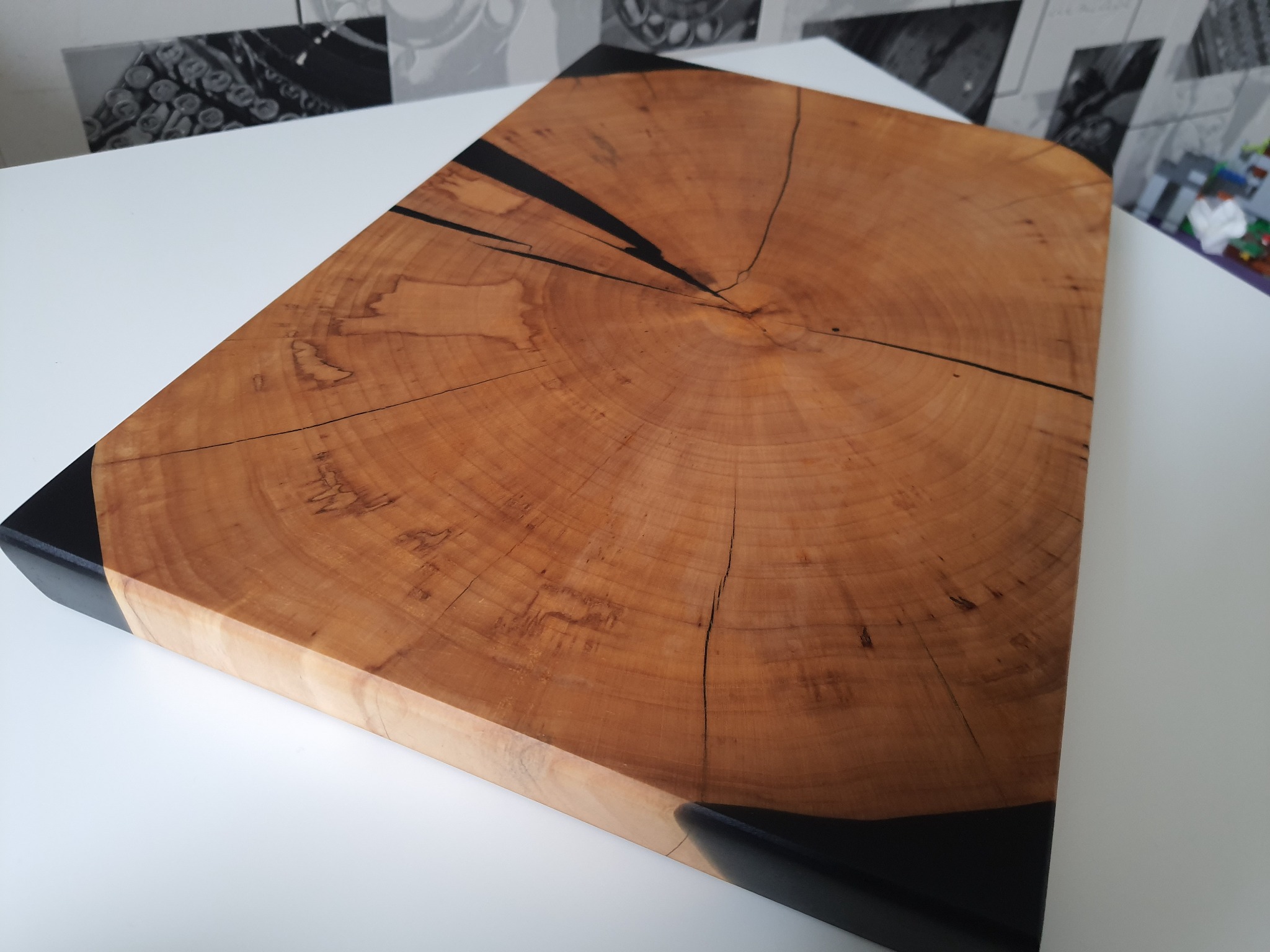 Kwadratowy blat z drewna z wyraźnym rysunkiem słojów, zalany czarną żywicą na krawędziach i w pęknięciach, leżący na białym stole.