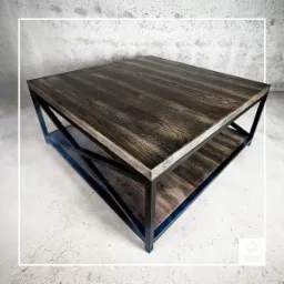 Kwadratowy stolik kawowy z ciemnego drewna z metalową ramą, dolna półka z drewna o podobnym odcieniu, surowy beton w tle.
