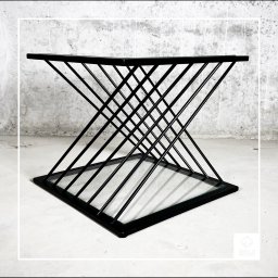 weld progress - Stolik kawowy z czarnych, metalowych prętów ułożonych w geometryczny wzór klepsydry z kwadratowym blatem z przezroczystego szkła, na tle surowego, betonowego muru.