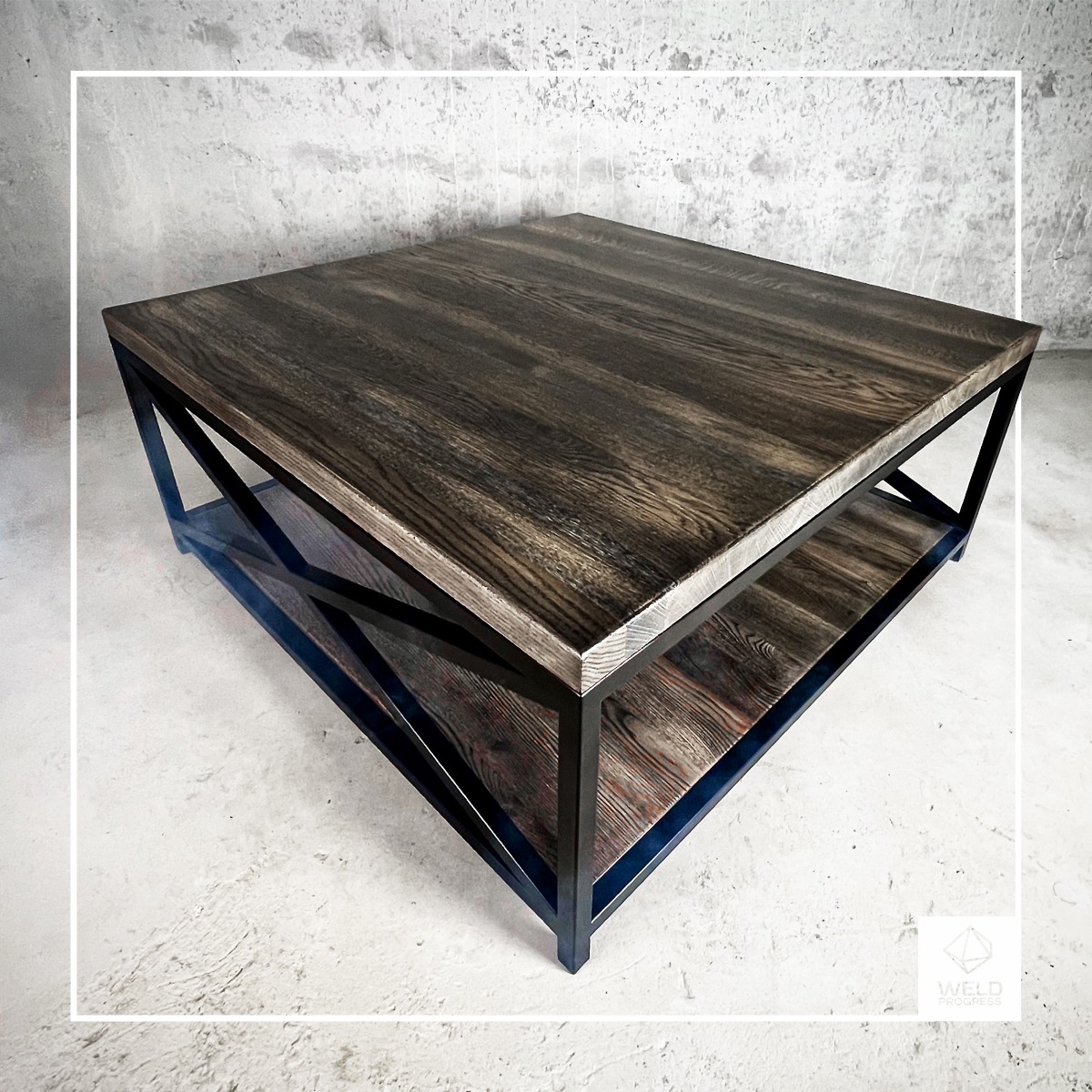 Kwadratowy stolik kawowy z ciemnego drewna z metalową ramą, dolna półka z drewna o podobnym odcieniu, surowy beton w tle.