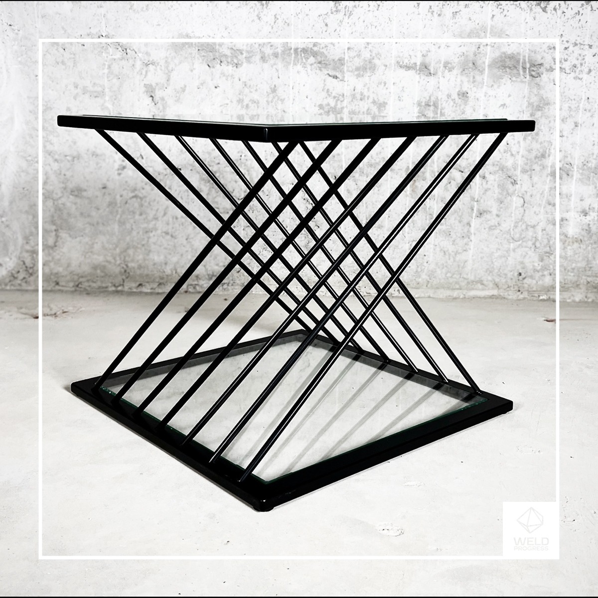 Stolik kawowy z czarnych, metalowych prętów ułożonych w geometryczny wzór klepsydry z kwadratowym blatem z przezroczystego szkła, na tle surowego, betonowego muru.