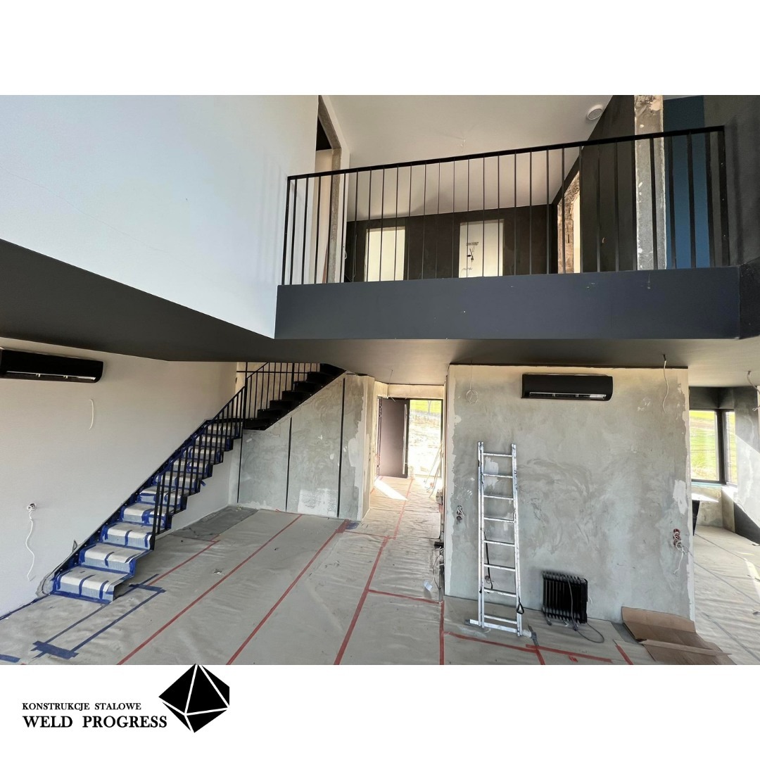 Wnętrze w trakcie wykańczania z zamontowanymi stalowymi schodami z czarną balustradą i czarnymi stopniami, zabezpieczonymi folią, widok na antresolę z czarną balustradą, surowe ściany, na podłodze...